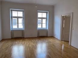 Pusty pokój z parkietem, białymi ścianami, dwoma oknami z grzejnikami pod spodem i otwartymi drzwiami do sąsiedniego pomieszczenia. Widoczne ślady po poprzednim malowaniu na ścianach.