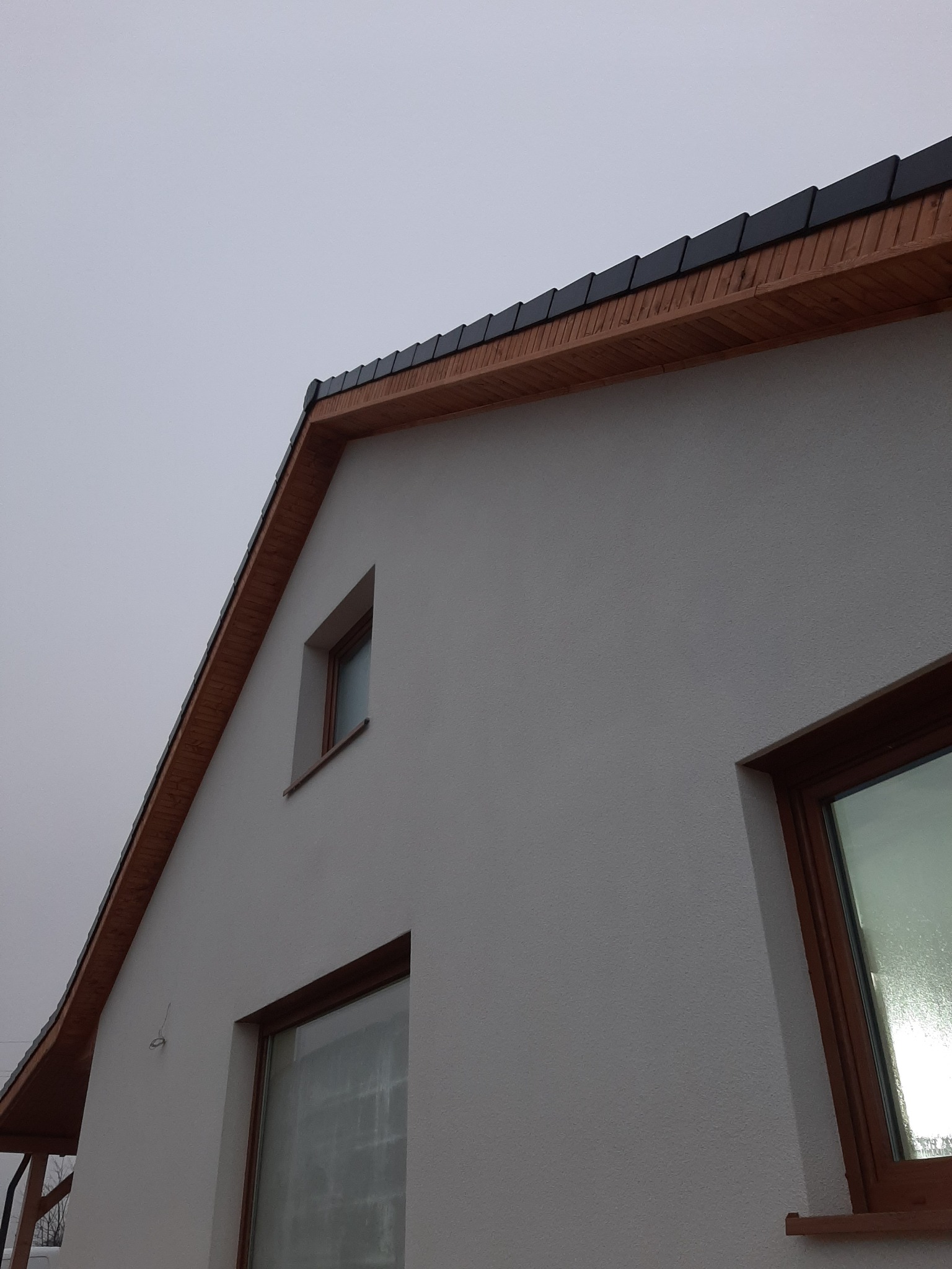 Fragment ściany szczytowej budynku z oknami i fragmentem dachu z drewnianą podbitką i czarną dachówką, widoczne niedociągnięcia w wykończeniu elewacji.