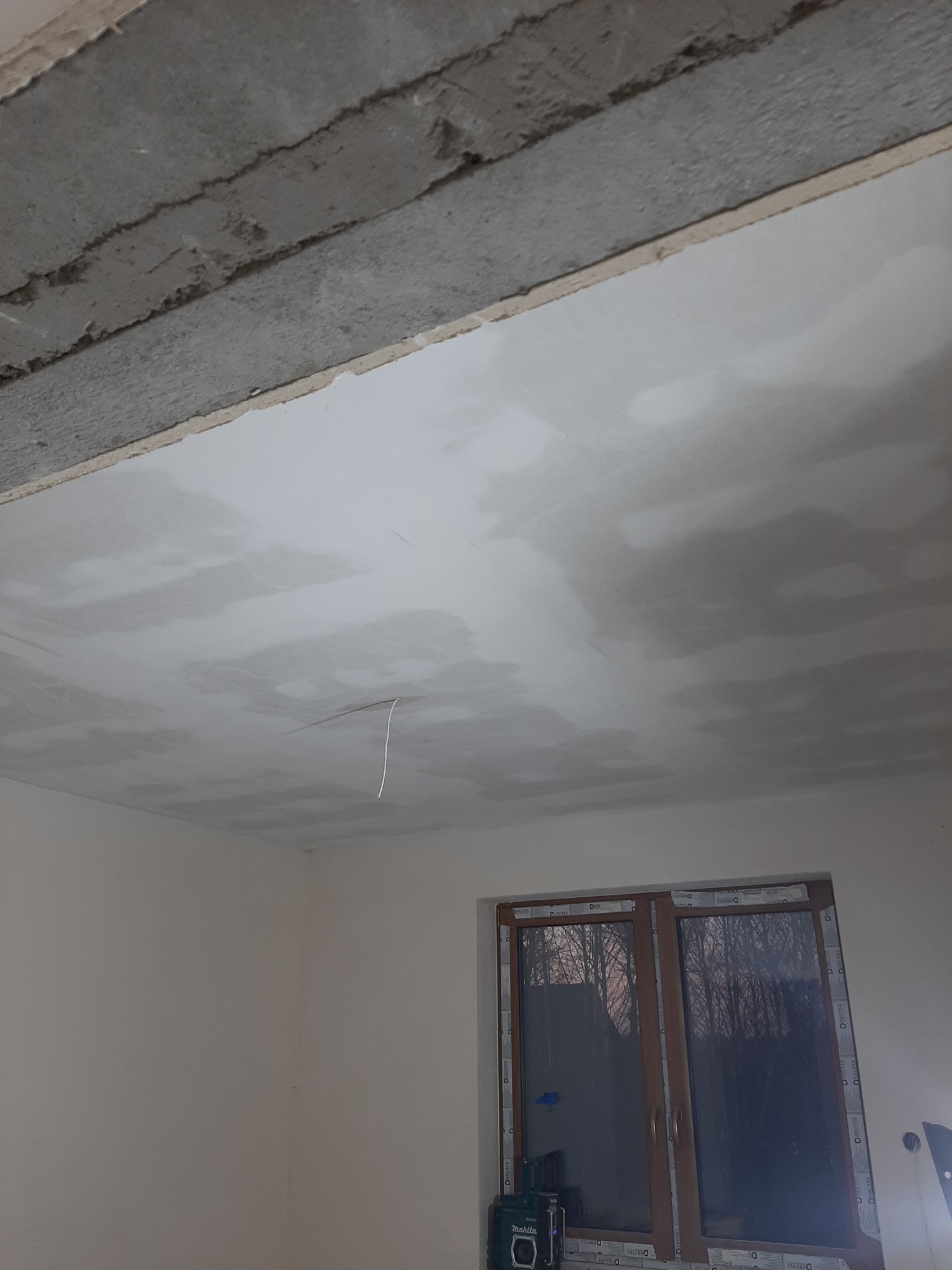 Niedokończony sufit po szpachlowaniu, widoczne nierówności i przygotowanie do dalszych prac wykończeniowych, z oknem w tle i narzędziami na podłodze.