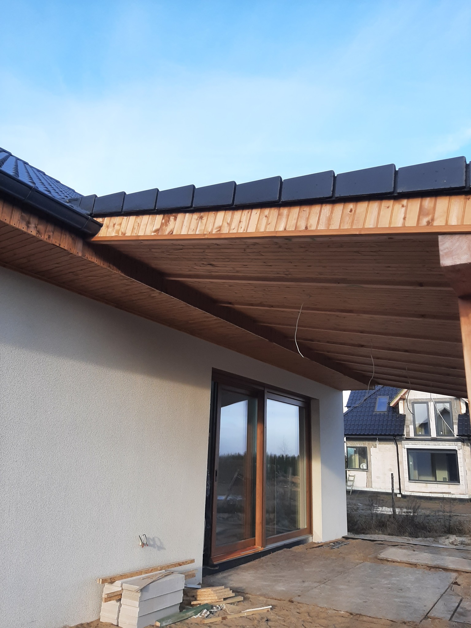 Zadaszenie tarasu z drewnianą konstrukcją i czarną dachówką, widoczne elementy elewacji z oknem przesuwnym, w tle budynek w trakcie budowy.