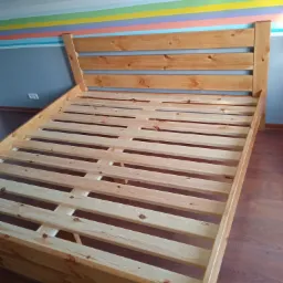 Drewniany stelaż łóżka z zagłówkiem, widoczny w pokoju z kolorowymi pasami na ścianie, bez materaca.