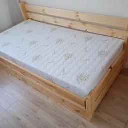 Drewniana rama łóżka z materacem z wzorem aloesu, widoczne słoje drewna i prosta konstrukcja.