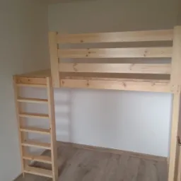 Drewniane łóżko na antresoli z drabinką w jasnym pokoju, widoczne połączenia śrubowe, surowe drewno, podłoga laminowana.