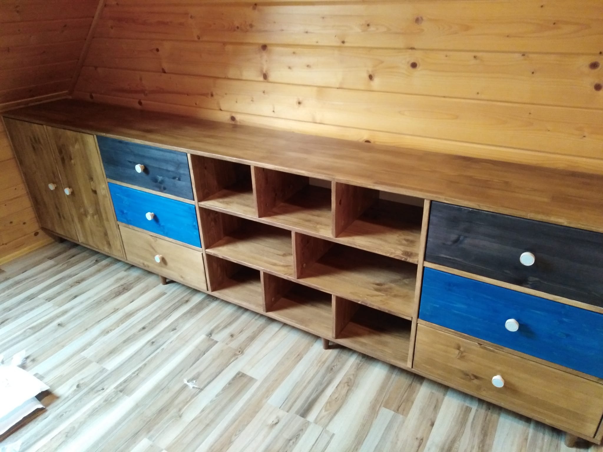 Drewniana szafa na wymiar z kolorowymi szufladami (niebieskie i grafitowe) w pomieszczeniu wykończonym boazerią, widoczne półki otwarte i gałki w kolorze białym.