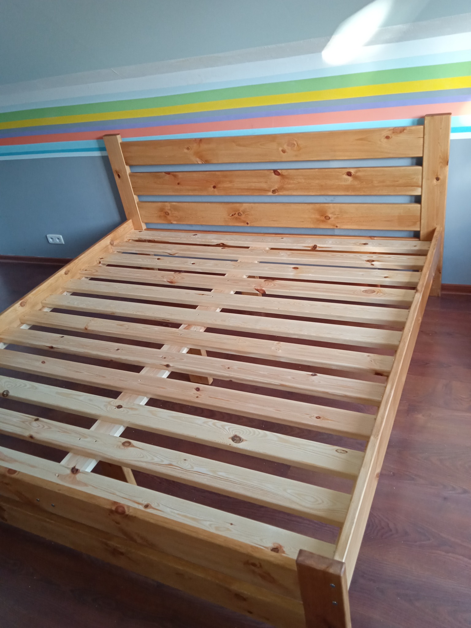 Drewniany stelaż łóżka z zagłówkiem, widoczny w pokoju z kolorowymi pasami na ścianie, bez materaca.