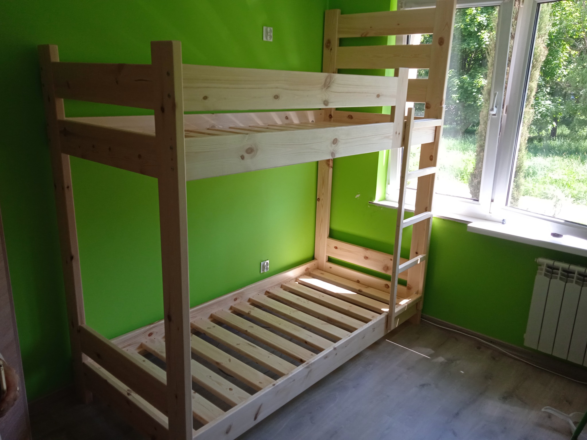 Surowa, sosnowa rama łóżka piętrowego w pokoju z zielonymi ścianami, widoczne drabinka i szczebelki, w tle okno z widokiem na zieleń.