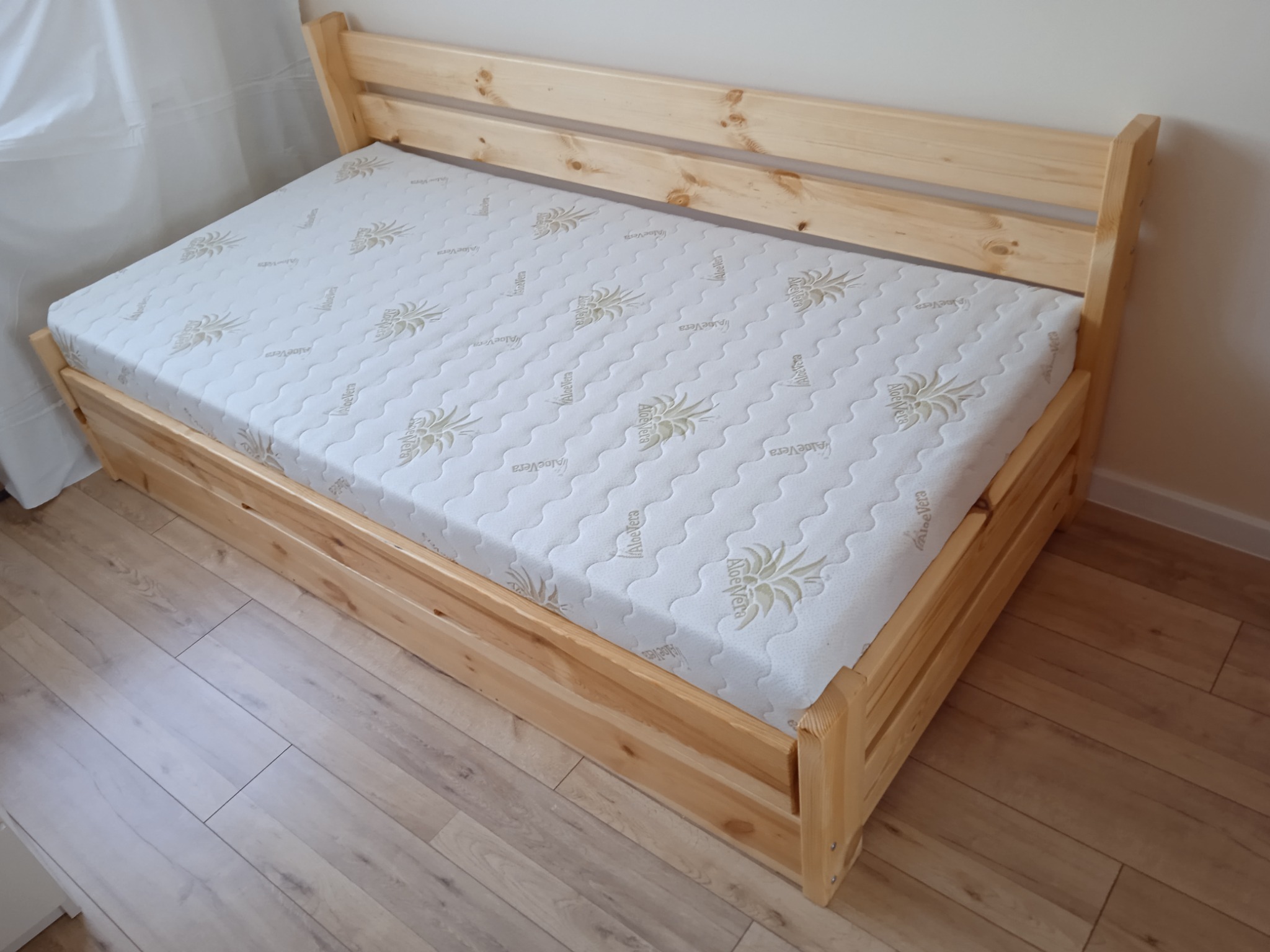 Drewniana rama łóżka z materacem z wzorem aloesu, widoczne słoje drewna i prosta konstrukcja.