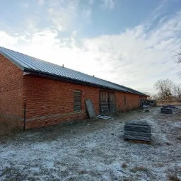Długi budynek z czerwonej cegły z nowym, metalowym pokryciem dachowym, widoczne palety z materiałami budowlanymi na ośnieżonym terenie.