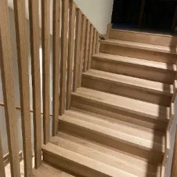 Drewniane schody z ażurową balustradą z pionowych listew, widok z dołu. Naturalne odcienie drewna i minimalistyczny design. Widoczny fragment okna.