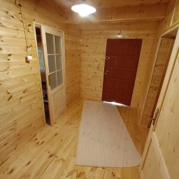 Usługi Stolarskie Mikołaj Adamczyk - Wnętrze wykończone boazerią: ściany, podłoga i sufit z jasnego drewna. Widoczne dwoje drzwi, jedno z przeszkleniami, oraz lampa sufitowa.