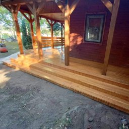 Usługi Stolarskie Mikołaj Adamczyk - Drewniany taras zadaszony w Gościeradowie Ukazowym. Solidna konstrukcja z jasnego drewna, trzy stopnie wejściowe. Widok na elementy konstrukcyjne dachu i ścianę budynku.