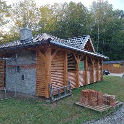 Usługi Stolarskie Mikołaj Adamczyk - Altana z kamienną obudową, drewnianymi lamelami i kominem w ogrodzie. Widoczne cegły i drabina sugerują etap budowy.
