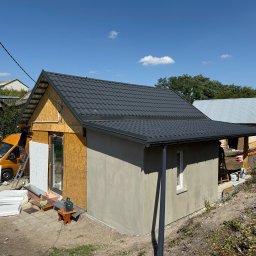 Usługi Stolarskie Mikołaj Adamczyk - Budowa małego domu: szara blachodachówka, ocieplanie ścian styropianem, żółty bus firmy w tle, drabina i narzędzia. Częściowo otynkowana ściana, okno.