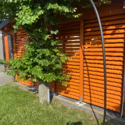 Pomarańczowa drewniana konstrukcja z żaluzjami osłonięta drzewem, widoczny fragment trawnika i chodnika z kostki. Konstrukcja z rynną i elementami montażowymi.