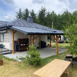 Nowoczesna wiata przy domu z szarym dachem blaszanym, drewnianymi słupami i meblami ogrodowymi. Widok na ogród, drzewa i zaparkowany bus.