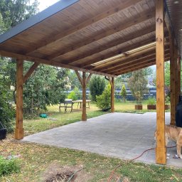 Usługi Stolarskie Mikołaj Adamczyk - Drewniana pergola z zadaszeniem w ogrodzie w Gościeradowie Ukazowym. Widoczne drewniane słupy, zadaszenie z desek oraz fragment betonowego tarasu i pies.
