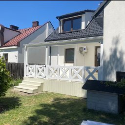 Usługi Stolarskie Mikołaj Adamczyk - Jasny taras z białą balustradą w kształcie krzyży, schodami i domkiem dla psa w ogrodzie. Widok na budynki mieszkalne w tle, słoneczny dzień.