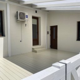 Usługi Stolarskie Mikołaj Adamczyk - Jasny, zadaszony taras z drewnianą podłogą i balustradą. Widoczne drzwi wejściowe, okna i klimatyzator. Oświetlenie dzienne, neutralne kolory.