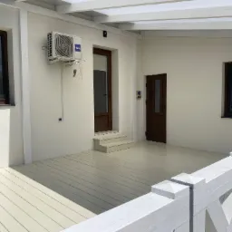 Jasny, zadaszony taras z drewnianą podłogą i balustradą. Widoczne drzwi wejściowe, okna i klimatyzator. Oświetlenie dzienne, neutralne kolory.