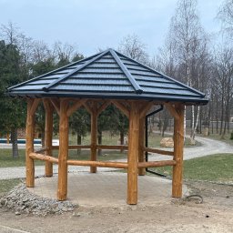 Usługi Stolarskie Mikołaj Adamczyk - Ośmioboczna altana z drewnianych bali, kryta ciemną dachówką, otoczona zielenią i kamieniami. Widok na ścieżkę i drzewa w tle. Naturalne oświetlenie.
