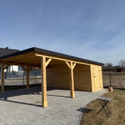 Usługi Stolarskie Mikołaj Adamczyk - Drewniana wiata garażowa z zamykanym pomieszczeniem gospodarczym, wykończona czarną blachą, na tle domu i błękitnego nieba. Mały stolik na trawie.