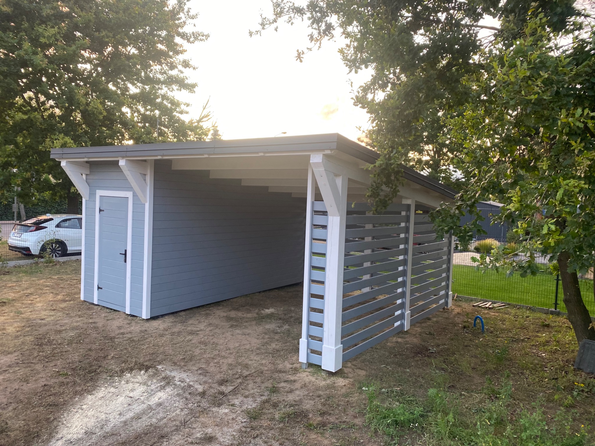 Drewniana altana z szarą zabudową i białymi elementami wykończeniowymi, zadaszona, z widocznymi poziomymi deskami po prawej stronie, na trawiastym terenie.