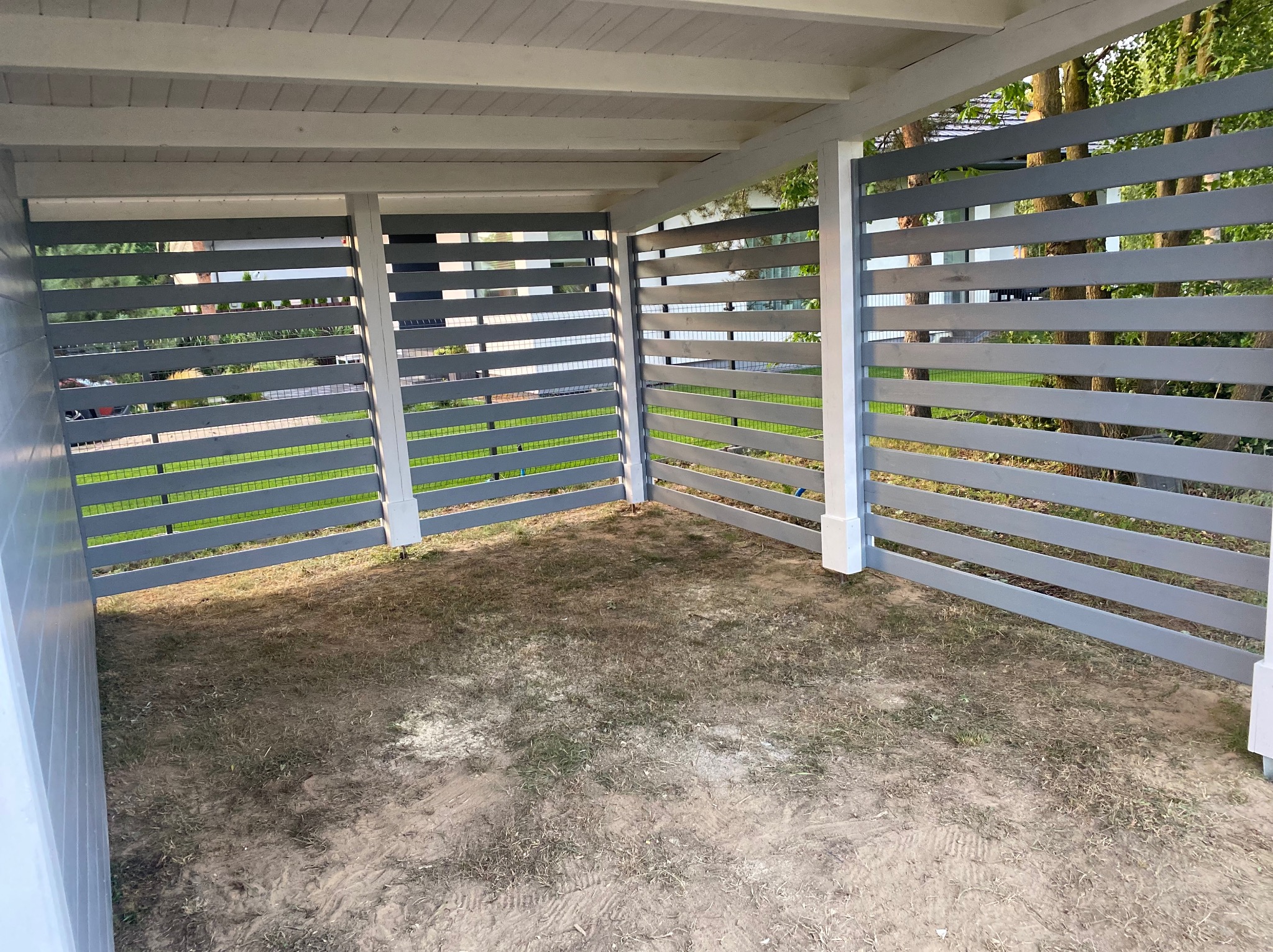 Szara, drewniana pergola z poziomymi listwami, widok z wnętrza, otoczenie zieleni. Konstrukcja z białymi słupkami, ziemia wewnątrz.