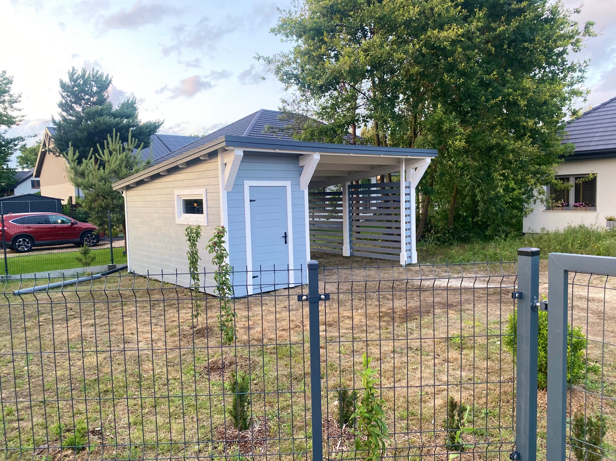 Niebieska altana z zadaszeniem na samochód, widoczna zza ogrodzenia. Budowla z oknem i drzwiami, zadaszenie wsparte na słupach. Tło stanowią domy i drzewa.