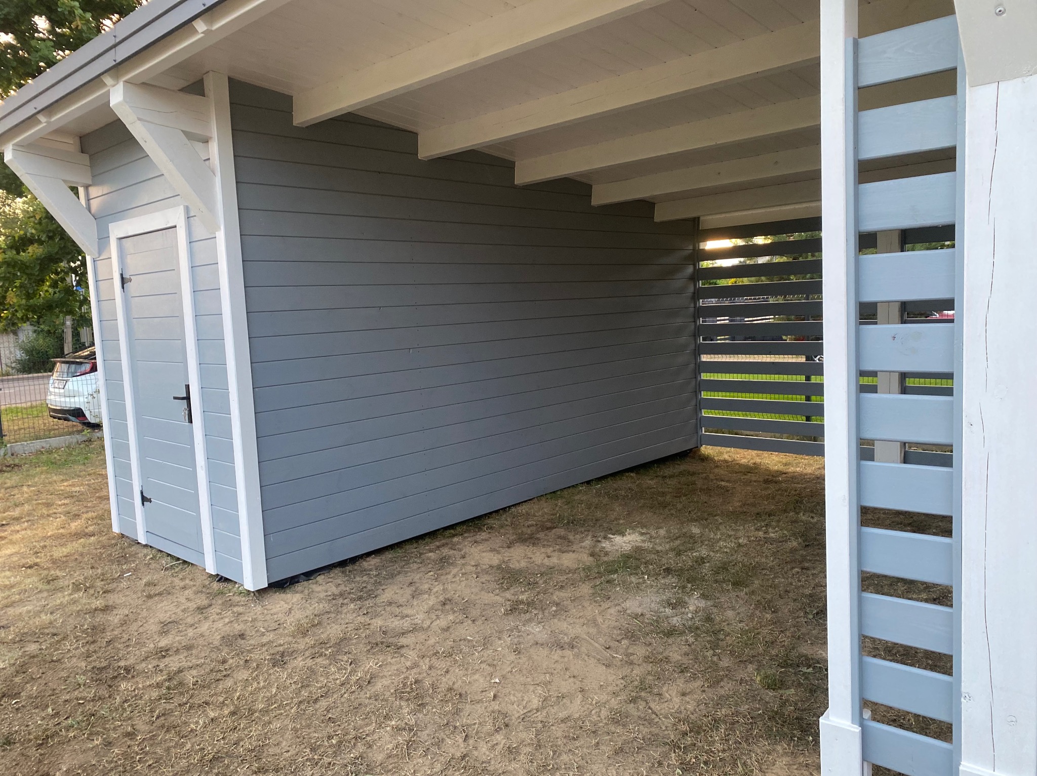 Szara altana z drewnianymi lamelami i białymi detalami, widok z zewnątrz, zarys drzew w tle. Konstrukcja na trawiastym podłożu.