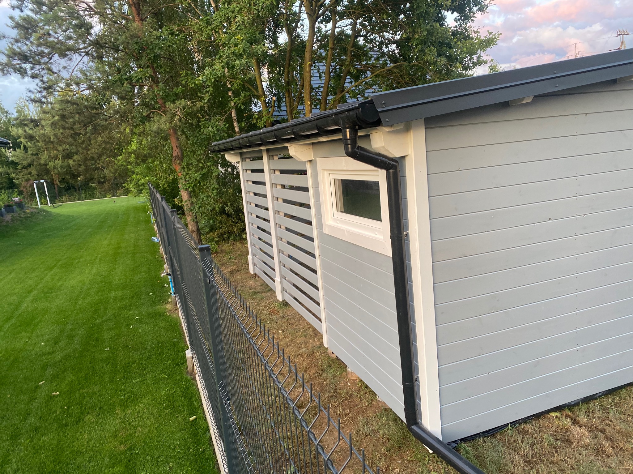 Szara, drewniana altana z oknem i czarną rynną, widziana zza metalowego ogrodzenia. Trawa i drzewa w tle, niebo z różowymi chmurami.