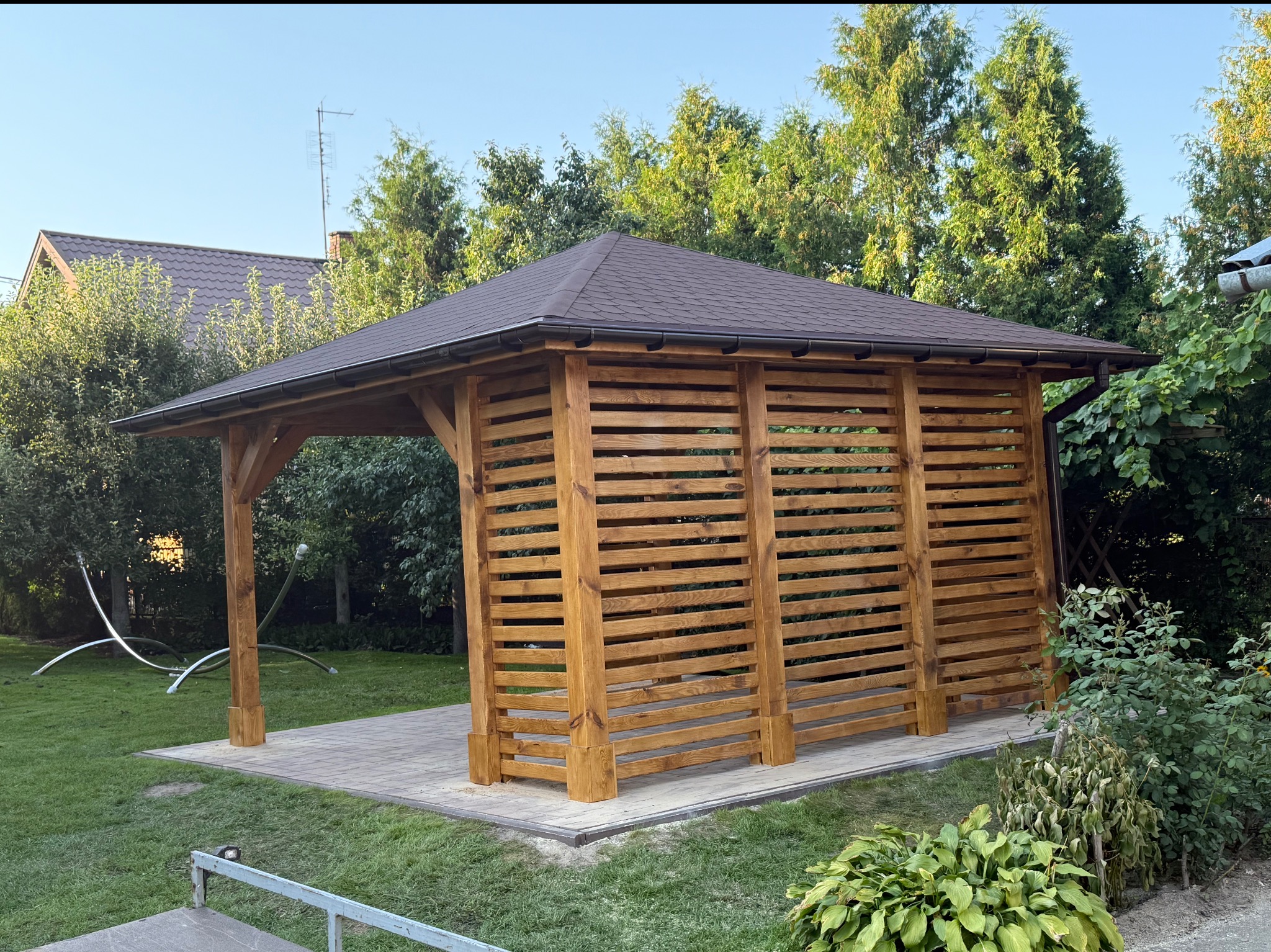Drewniana altana z brązowym dachem na kostce brukowej, otoczona zielenią. Konstrukcja z poziomych desek, widoczne drzewa w tle i fragment domu. Słoneczny dzień.