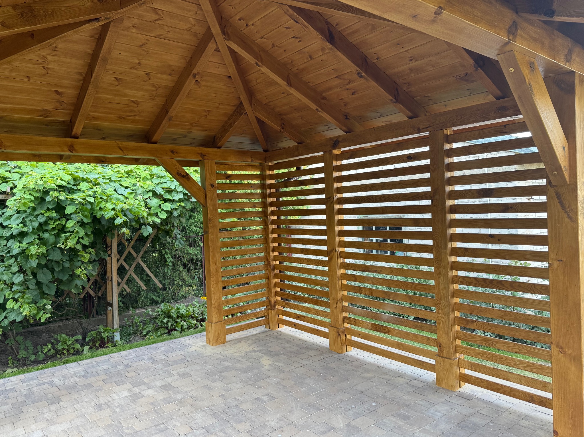 Drewniana altana z ażurowymi ścianami, widok z wnętrza na kostkę brukową i zieleń. Konstrukcja z belek, widoczny fragment dachu. Ujęcie z poziomu podłoża.