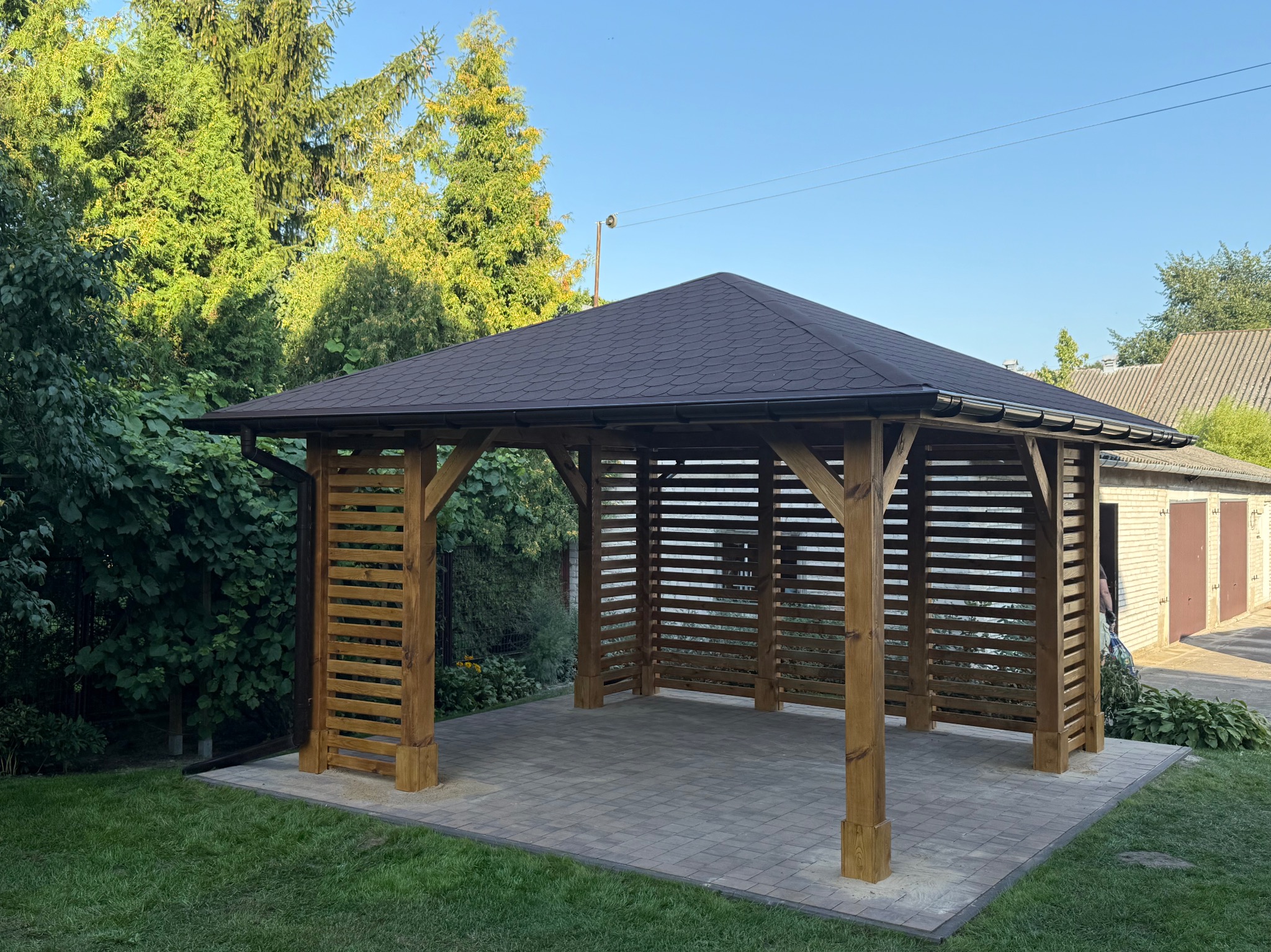 Drewniana altana ogrodowa z dachem krytym gontem bitumicznym, otoczona zielenią. Konstrukcja z poziomych listew zapewnia częściową osłonę, stoi na kostce brukowej.