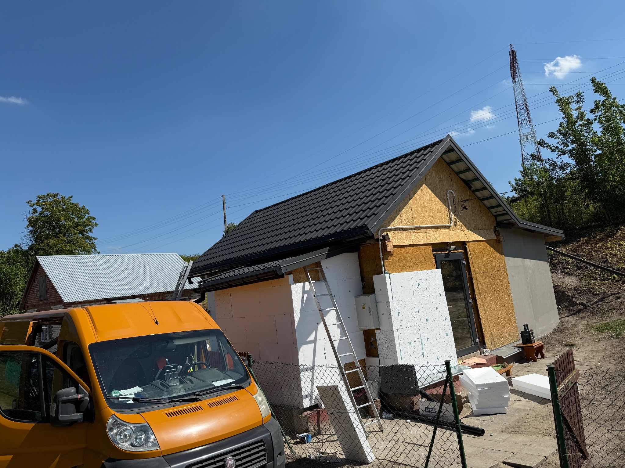 Budynek w trakcie ocieplania, z widocznym fragmentem dachu pokrytego czarną dachówką, białym styropianem, drabina i pomarańczowy bus transportowy.