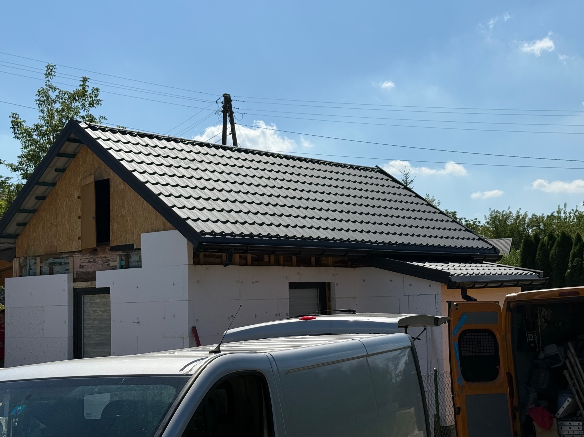 Dom w trakcie budowy z czarnym dachem i ociepleniem styropianem, widoczne busy transportowe. Budowa w toku, z elementami konstrukcyjnymi bez wykończenia.