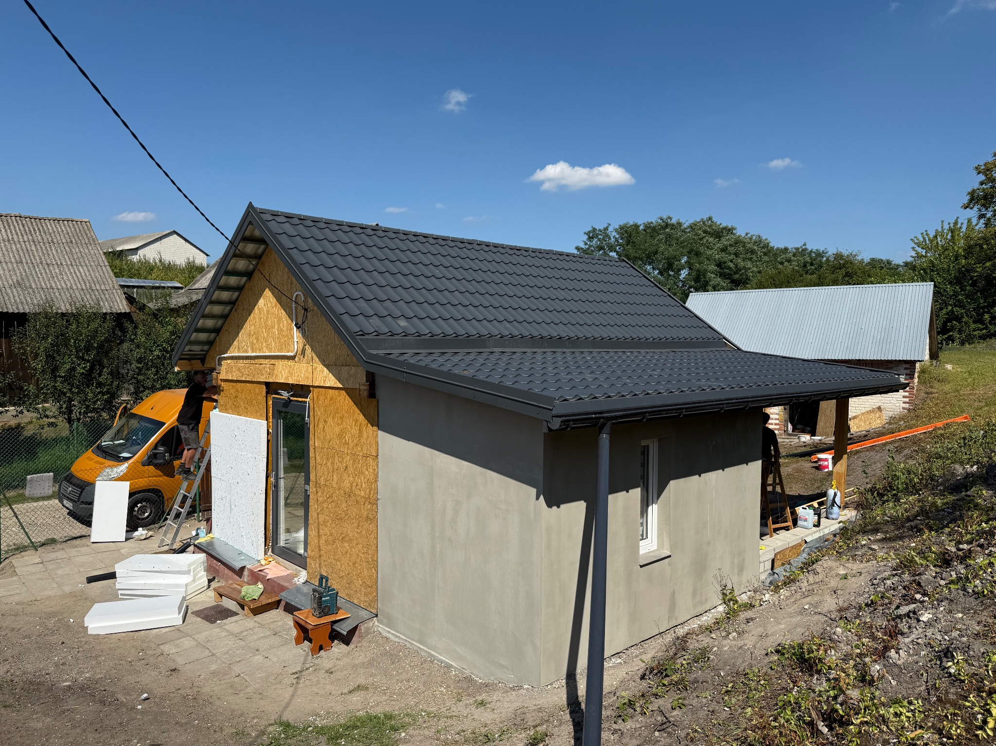 Budowa małego domu: szara blachodachówka, ocieplanie ścian styropianem, żółty bus firmy w tle, drabina i narzędzia. Częściowo otynkowana ściana, okno.