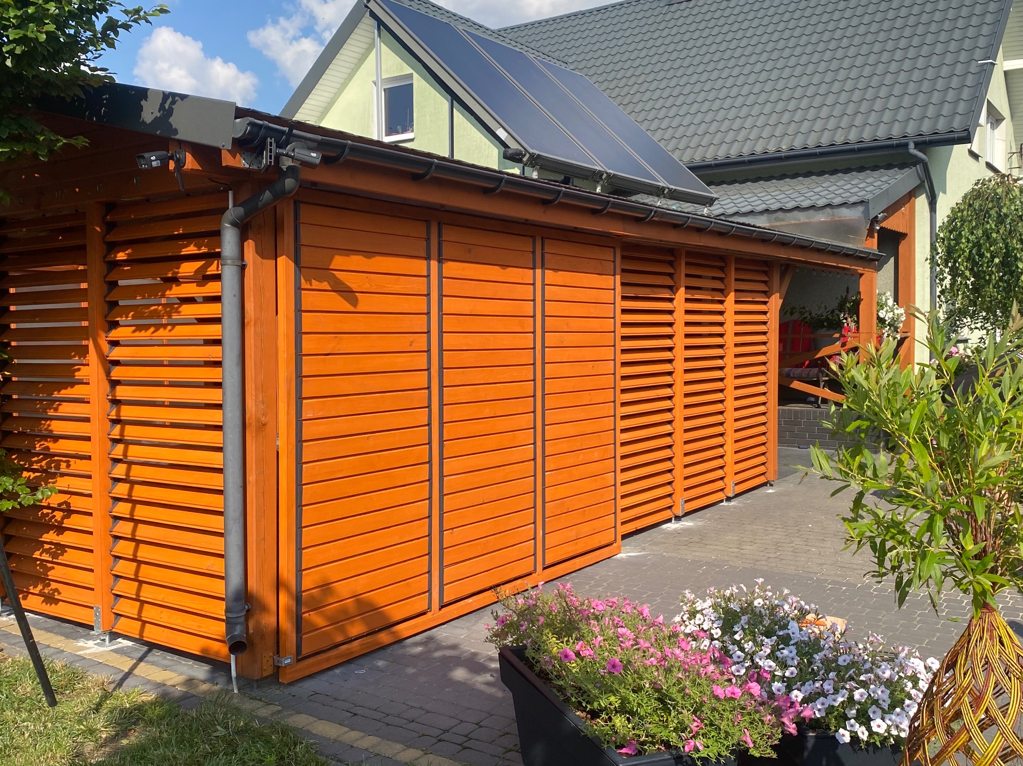 Pomarańczowa pergola z drewnianymi żaluzjami, zadaszona, z rynną, na tle domu z panelami słonecznymi. W tle kwiaty w donicy, kostka brukowa.