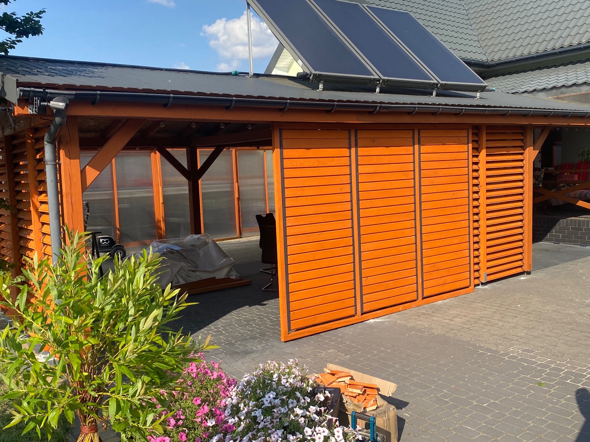 Drewniana altana z przesuwnymi panelami w kolorze pomarańczowym, z panelami solarnymi na dachu. Widoczne kwiaty i kostka brukowa.