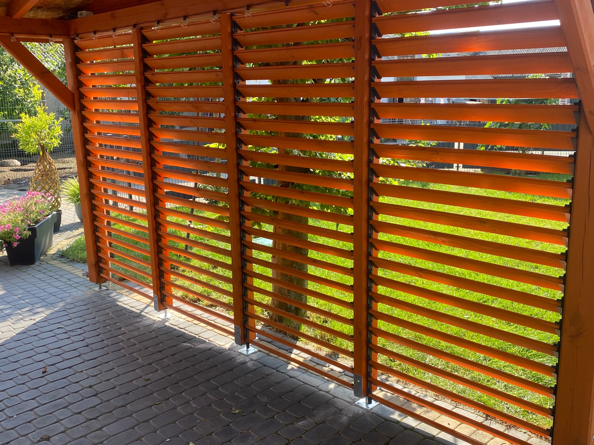 Drewniana pergola z żaluzjowymi panelami w ogrodzie, widok z zewnątrz. Widoczne elementy konstrukcyjne, kostka brukowa i zieleń w tle. Ciepłe oświetlenie.