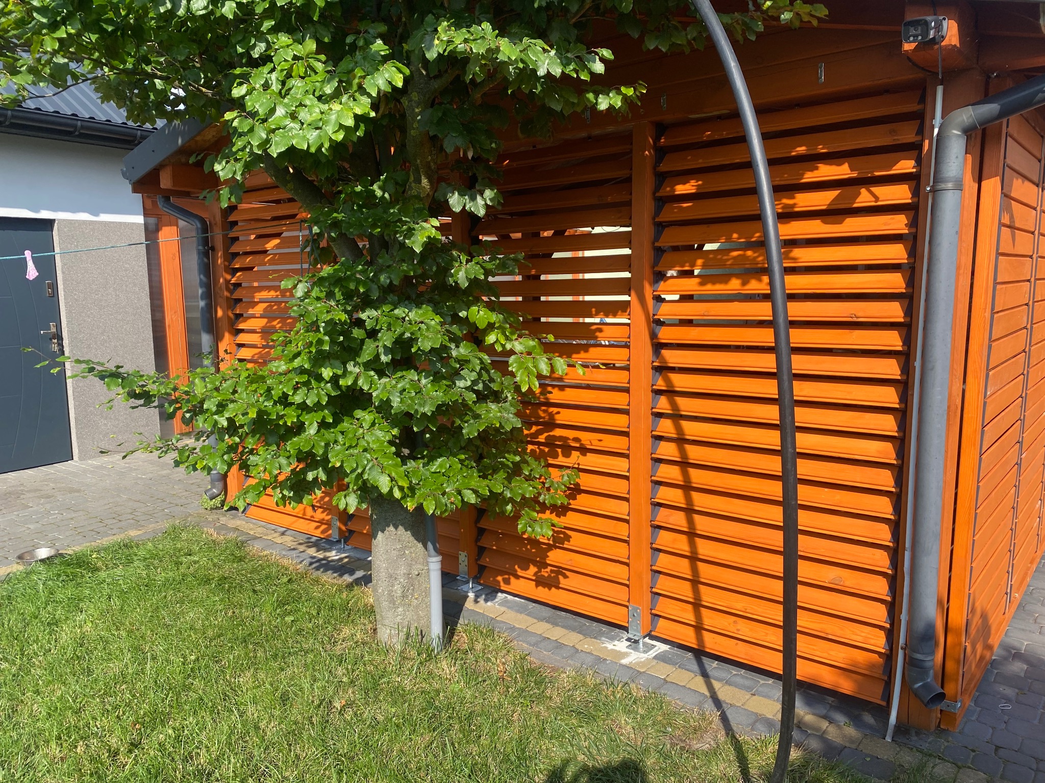 Pomarańczowa drewniana konstrukcja z żaluzjami osłonięta drzewem, widoczny fragment trawnika i chodnika z kostki. Konstrukcja z rynną i elementami montażowymi.