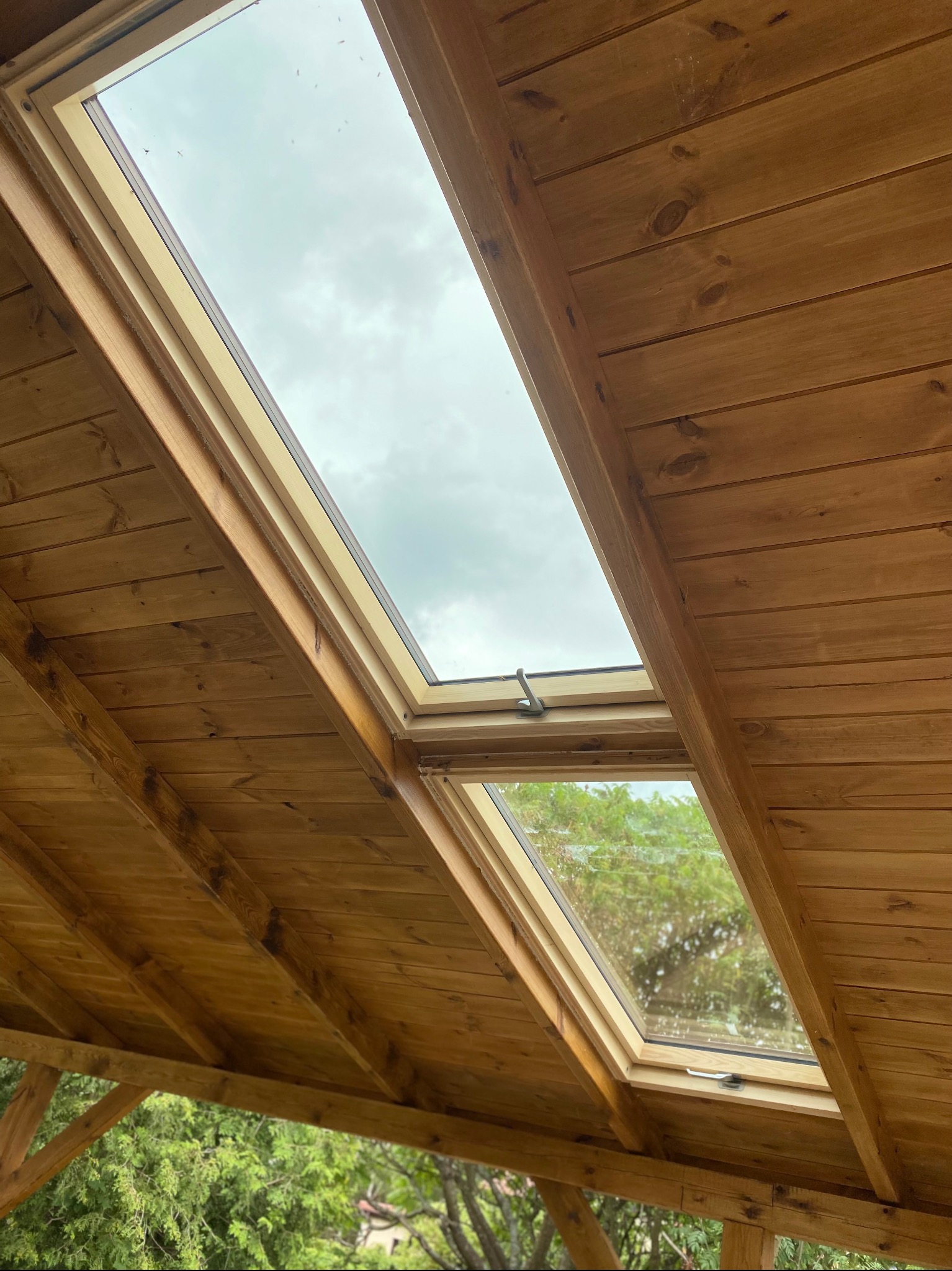 Dwa okna dachowe w drewnianej konstrukcji, widok z dołu. Widoczne niebo i zieleń drzew przez okna. Naturalne światło. Konstrukcja z desek o ciepłym odcieniu brązu.