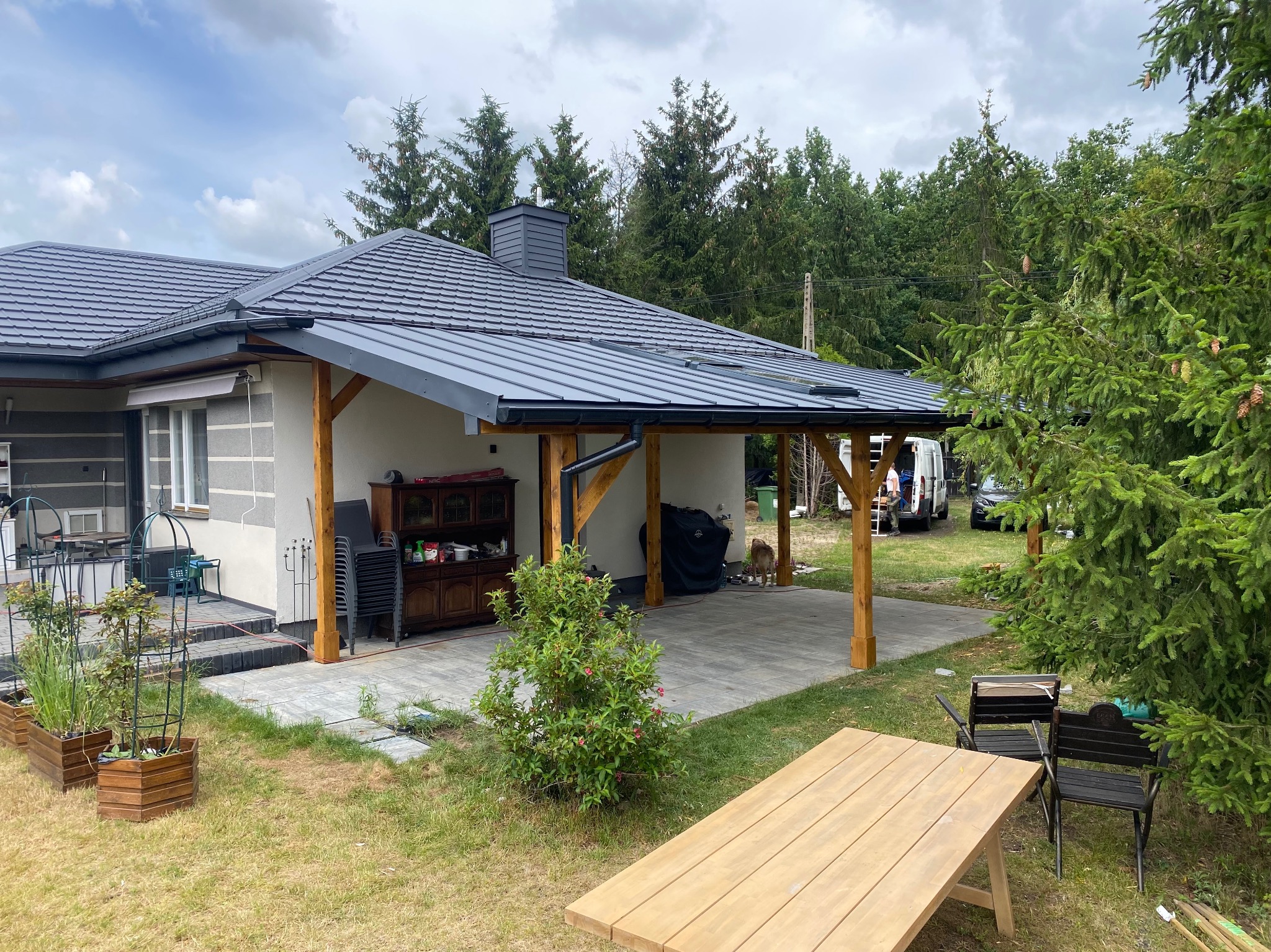 Nowoczesna wiata przy domu z szarym dachem blaszanym, drewnianymi słupami i meblami ogrodowymi. Widok na ogród, drzewa i zaparkowany bus.