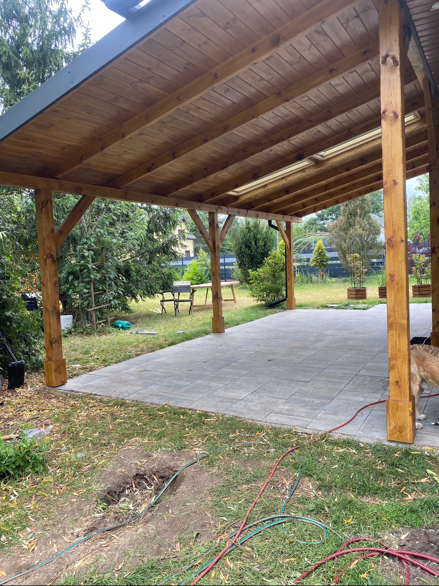 Drewniana pergola z zadaszeniem z desek i szarą obróbką blacharską, na betonowym tarasie. W tle zieleń ogrodu.