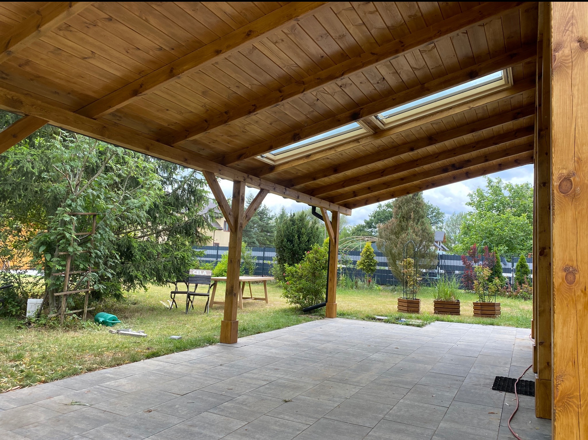 Drewniana pergola z oknami dachowymi, widok z zewnątrz na ogród z meblami i roślinami. Solidna konstrukcja, naturalne oświetlenie, estetyczne wykończenie.