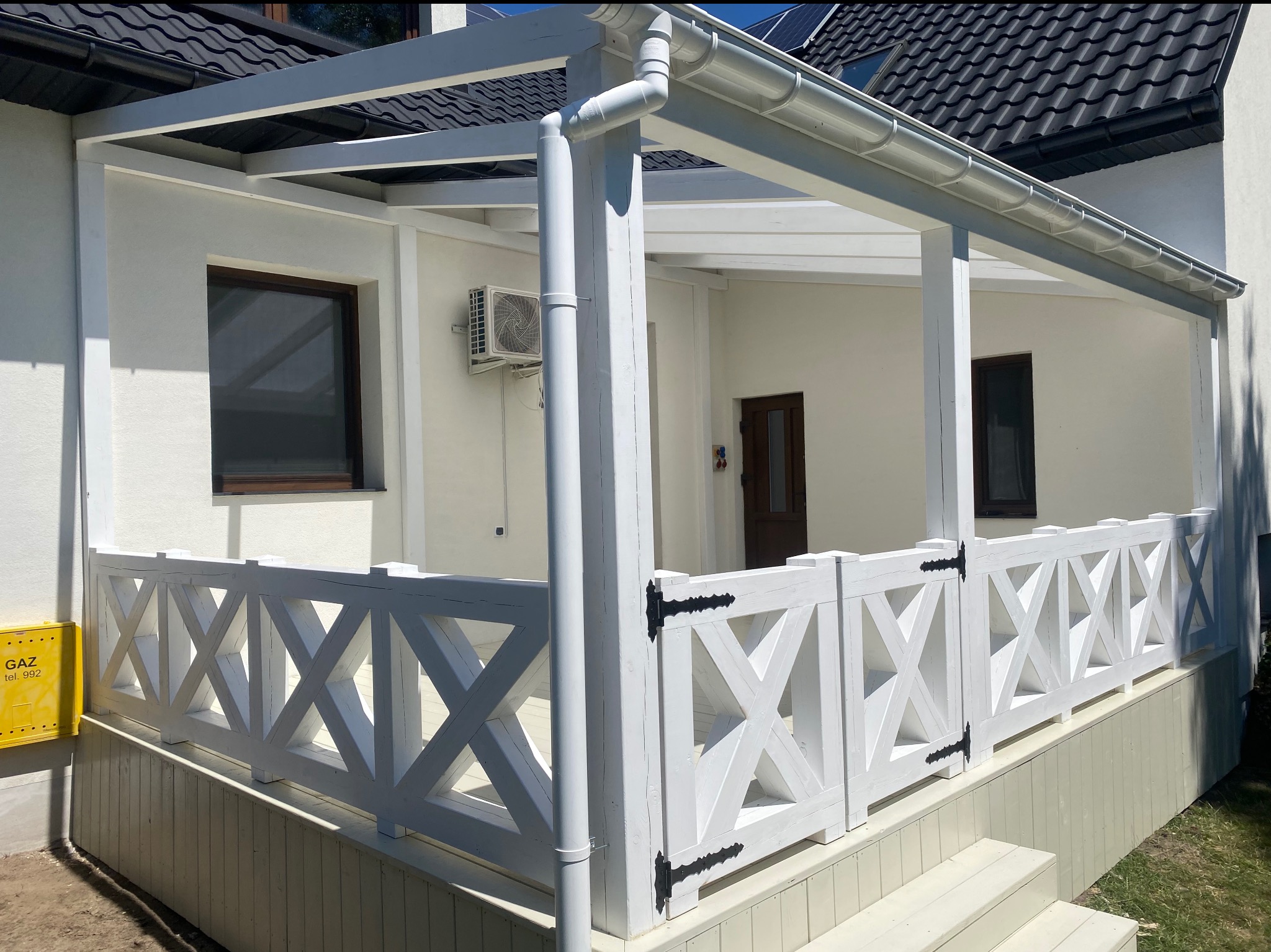 Biała pergola z drewnianą balustradą w kształcie krzyżyków, z widocznym klimatyzatorem na ścianie budynku i fragmentem czarnego dachu w tle.