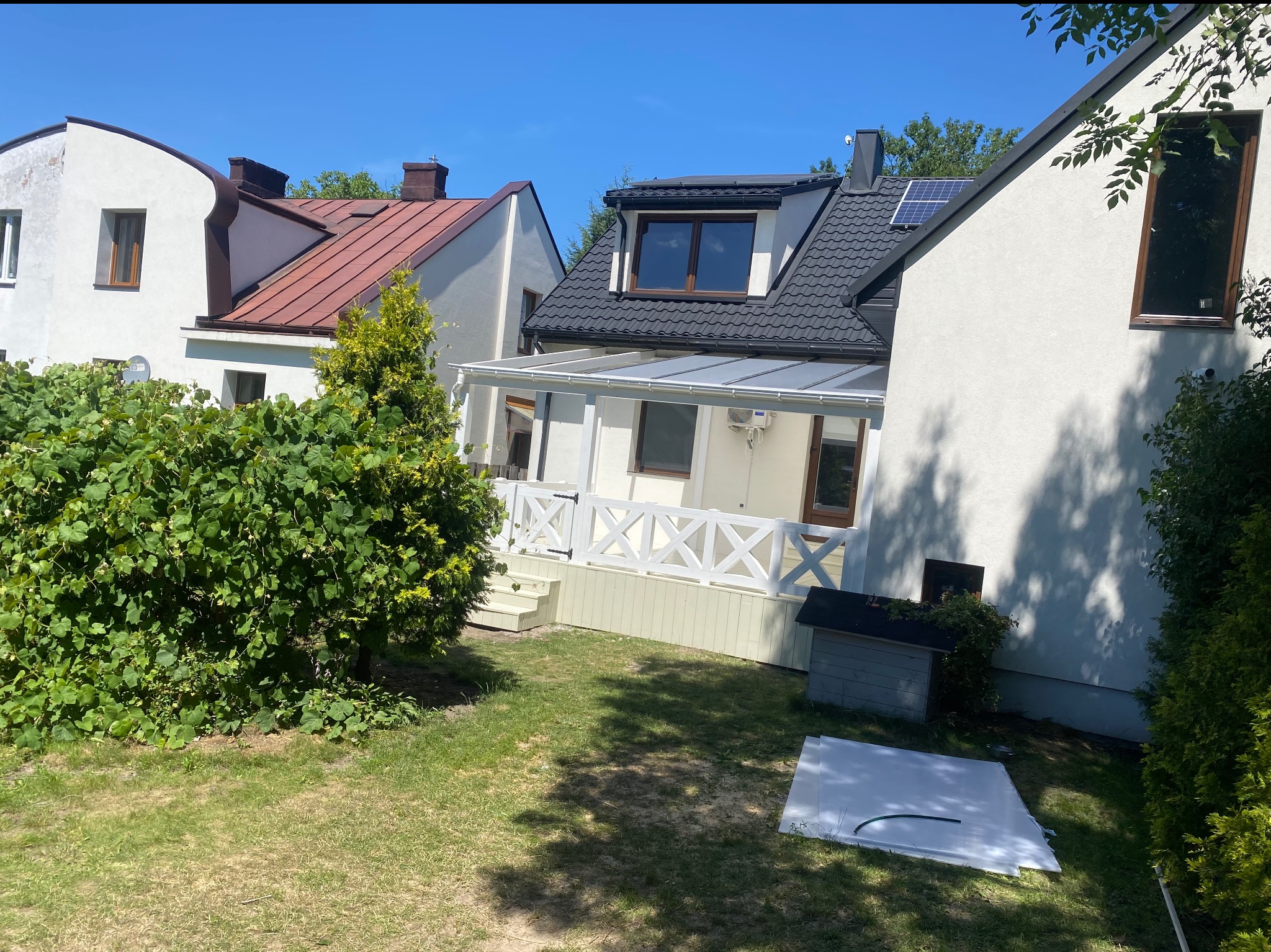 Biały dom z czarnym dachem i panelem słonecznym, widok z ogrodu na taras z białą balustradą i przezroczystym zadaszeniem, w tle sąsiednie domy.