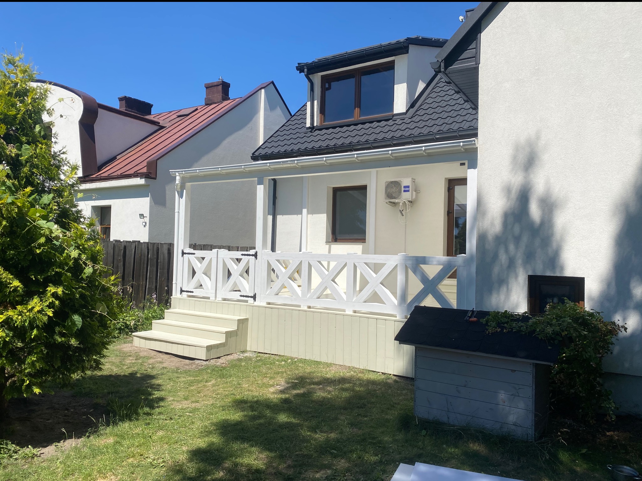 Jasny taras z białą balustradą w kształcie krzyży, schodami i domkiem dla psa w ogrodzie. Widok na budynki mieszkalne w tle, słoneczny dzień.