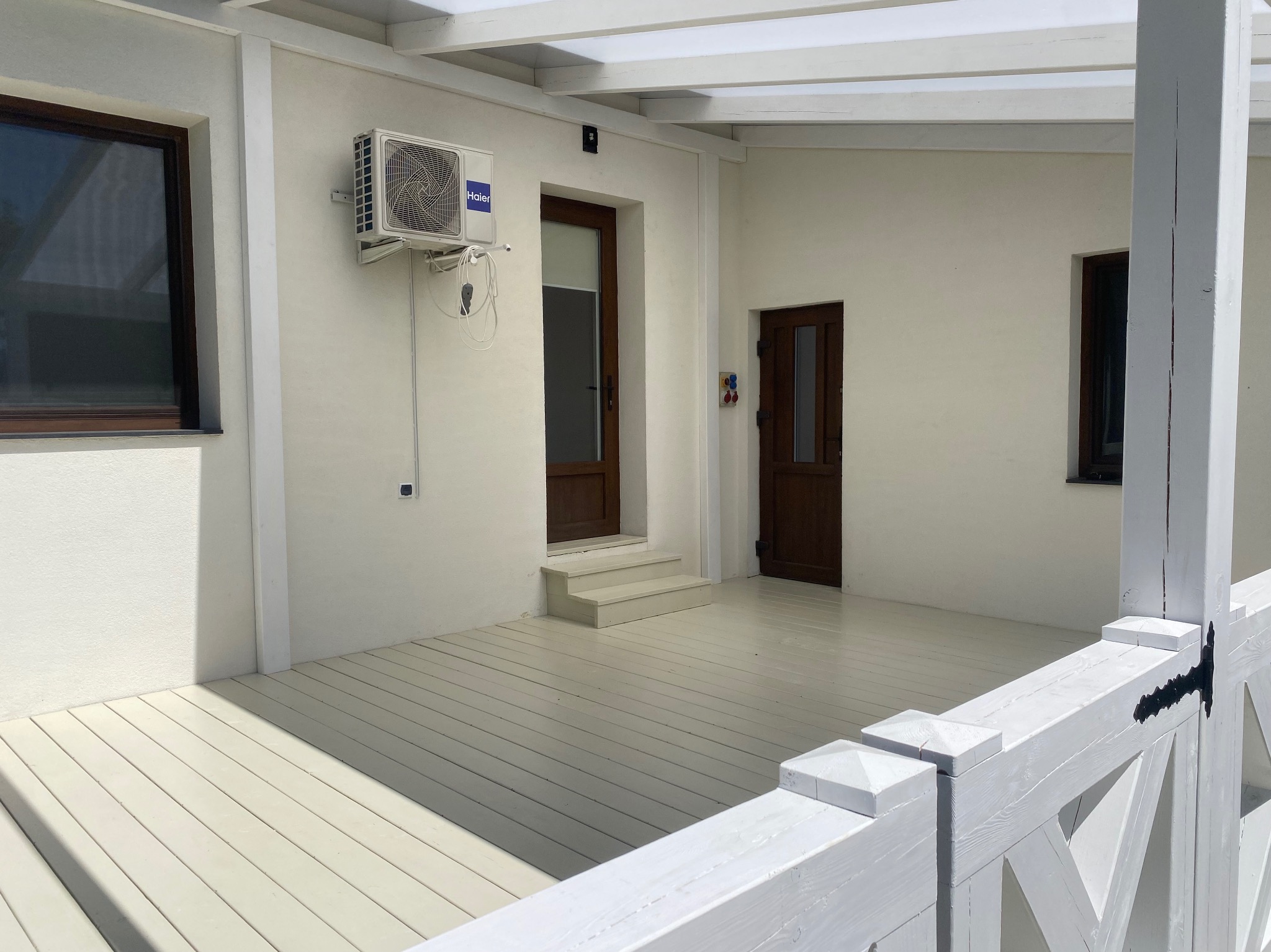 Jasny, zadaszony taras z drewnianą podłogą i balustradą. Widoczne drzwi wejściowe, okna i klimatyzator. Oświetlenie dzienne, neutralne kolory.