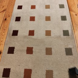 Budowanko i Sprzątanko Renata Grześkowiak - Beżowy dywan w kwadraty w odcieniach brązu i zieleni, częściowo wyczyszczony, leżący na drewnianej podłodze, z widocznym fragmentem odkurzacza piorącego.