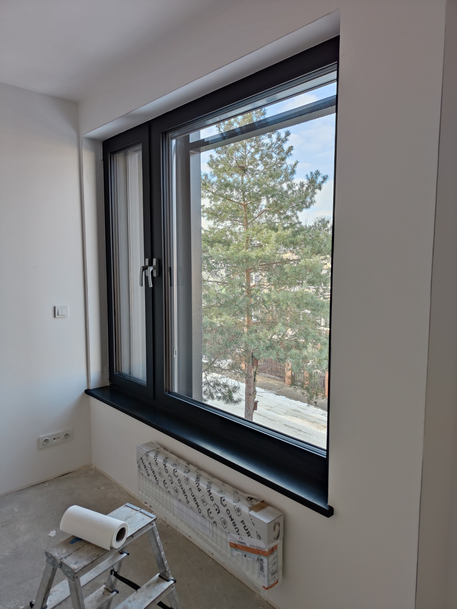 Wnętrze pokoju w trakcie remontu z czarnym oknem, widok na sosnę za oknem, drabina malarska z rolką papieru i grzejnik pod oknem jeszcze w opakowaniu.